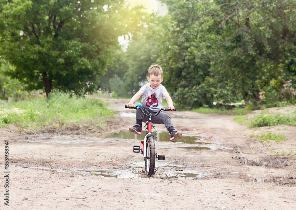 Naklejka premium 5 years boy is riding a bicycle through puddles in village in summer