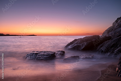 A Sandy Beach and Warm Sunset in Newquay, Cornwall, UK, with foreground rock formation and misty, long exposure waves. Dark mode style background.
