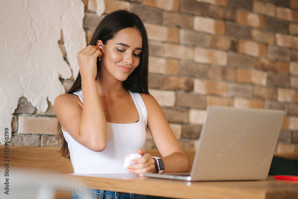 Obraz premium Attractive young girl use wireless headphones and working on laptop. Woman in white t-shirt and denim. Student study in cafe. Alone in cafe