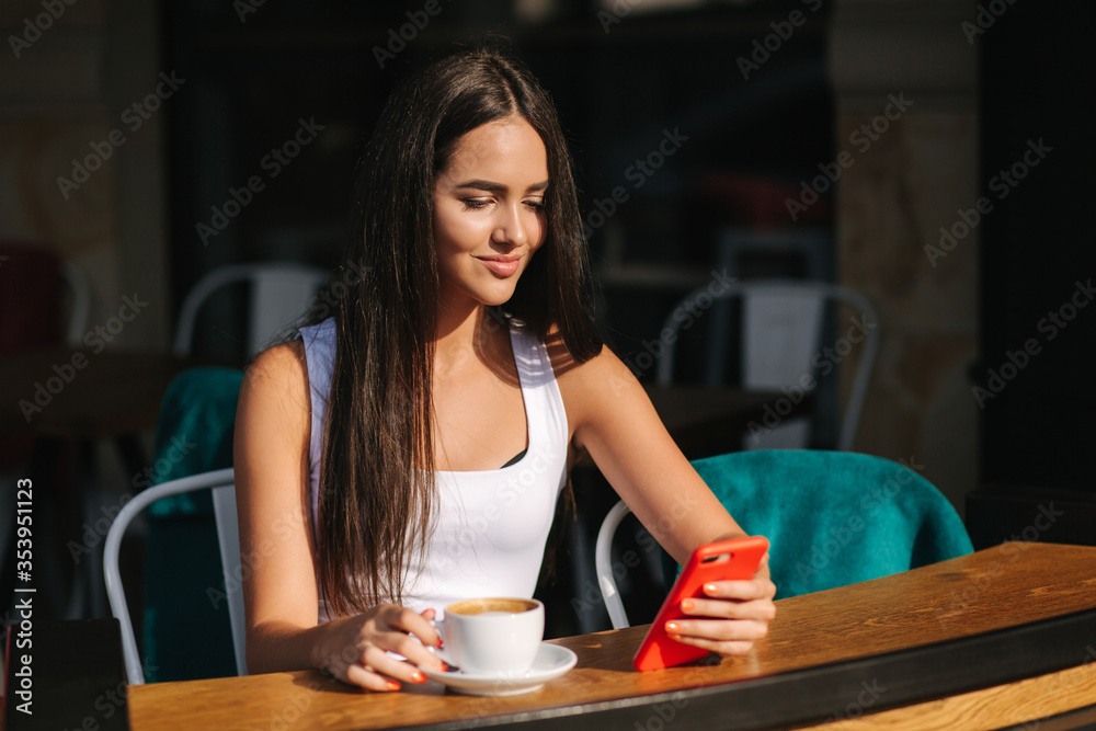 Attractive young woman using phone in cafe on terrace. Social distancing. Beautiful brunette girl chatting and drink coffee Attractive young woman using phone in cafe on terrace. Social distancing. Beautiful brunette girl chatting and drink coffee