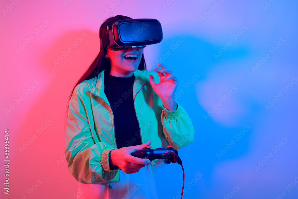 Laughing smiling surprise girl in neon with VR glasses holds a gamepad in her hands and plays games. Virtual world, technology. Laughing smiling surprise girl in neon with VR glasses holds a gamepad in her hands and plays games. Virtual world, technology.