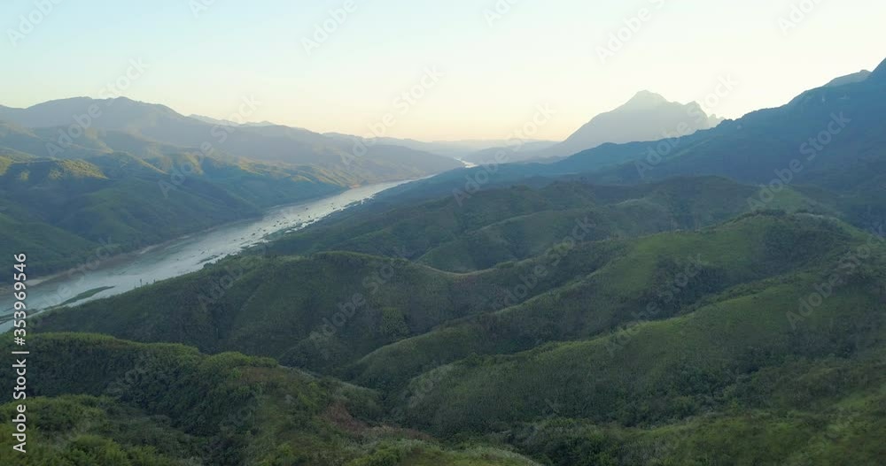 Aerial shot of river amidst green mountains during sunrise, drone flying forward over trees in lush forest against sky - Luang Prabang, Laos