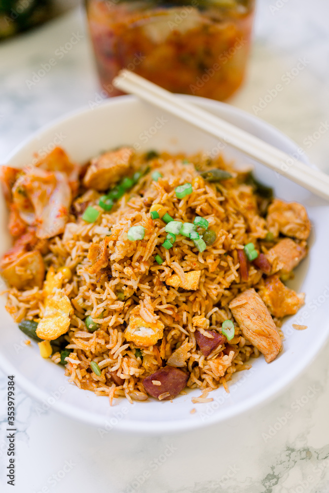 Delicious Chicken Fried Rice with Egg and a Side of Kimchi Served in a White Bowl. A package of Kimchi is sitting in the background. Chopsticks rest on the bowl. Delicious Chicken Fried Rice with Egg and a Side of Kimchi Served in a White Bowl. A package of Kimchi is sitting in the background. Chopsticks rest on the bowl.