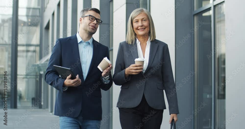Male and female business partners walking the street together with coffee to-go in hands and talking cheerfully. Businesswoman and businessman going in the morning to work and discussing job. Male and female business partners walking the street together with coffee to-go in hands and talking cheerfully. Businesswoman and businessman going in the morning to work and discussing job.
