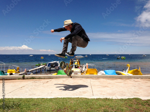 Young White Man in Dark Clothes and Beige Hat Doing a Skateboard Trick by the Titikaka Lake with Boats and Water Toys in Background in Copacabana, La Paz / Bolivia