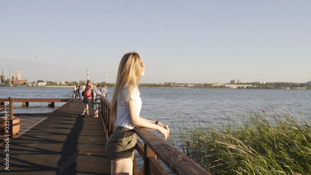 Happy smiling young blond woman in trendy sunglasses looking at summer sunset on river. Cute face model smiling on cityscape lake background. Girl looks joy at camera. Blond hair fly fluttering wind.