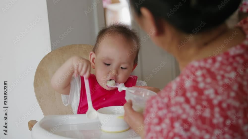 Close up of asian baby girl eating grandmother feeding baby girl with spoon at home Close up of asian baby girl eating grandmother feeding baby girl with spoon at home