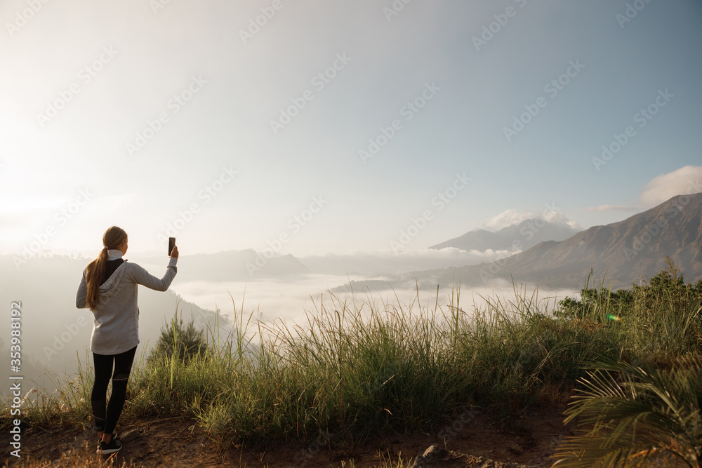 Young lady hiker with backpack standing on top of the mountain and taking a picture of valley with view on Batur vo;cano high in the mountains. Young female is shooting video on cell telephone Young lady hiker with backpack standing on top of the mountain and taking a picture of valley with view on Batur vo;cano high in the mountains. Young female is shooting video on cell telephone