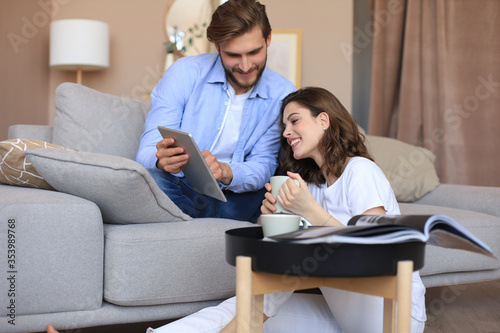 Happy young couple sitting, relaxing on floor in living room watching media content online, doing online shopping in a tablet.