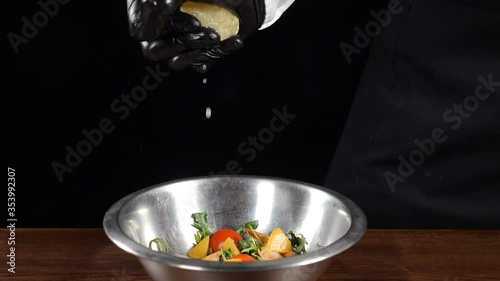 Close-up of chef hand in black gloves, squeezing fresh lemon into steel bowl with fresh salad. Shot on black background. Splashes and drops in slow motion. Full hd