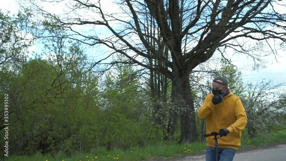 pensioner in yellow hoodie facial mask and gloves rides scooter along village road past spring trees slow motion closeup pensioner in yellow hoodie facial mask and gloves rides scooter along village road past spring trees slow motion closeup