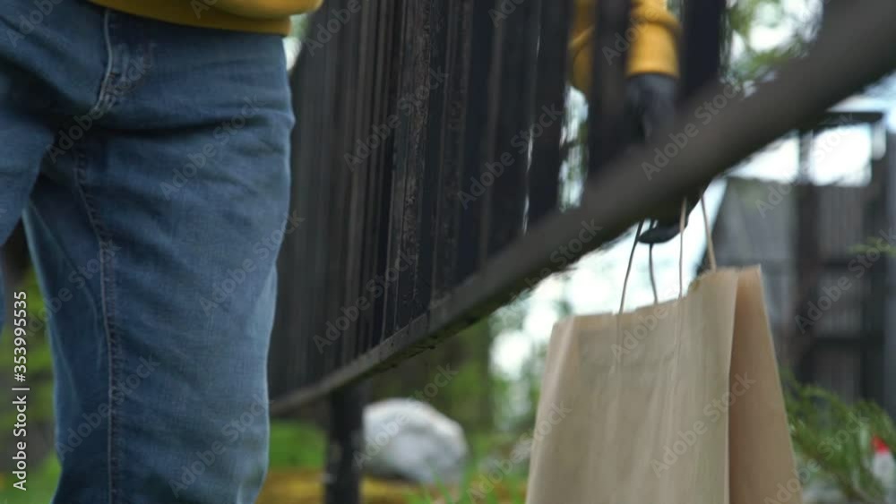 man in jeans takes large paper bag from behind black metal fence among grass and flowers slow motion close low angle shot