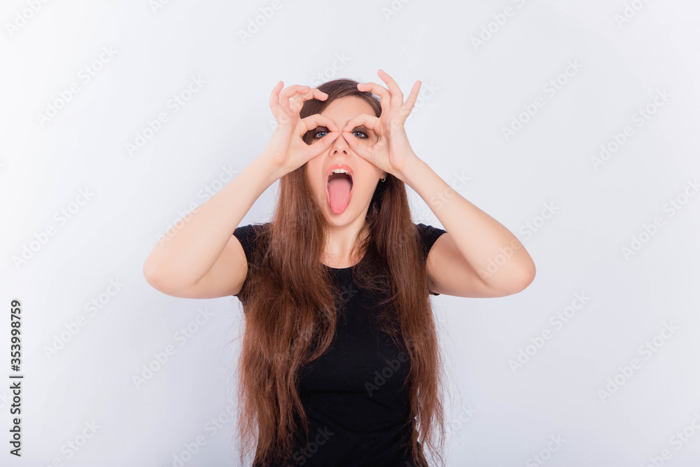 Girl in a black T-shirt with long hair grimaces and shows glasses with her hands isolated on a white background. The concept of an emotional portrait. Girl in a black T-shirt with long hair grimaces and shows glasses with her hands isolated on a white background. The concept of an emotional portrait.