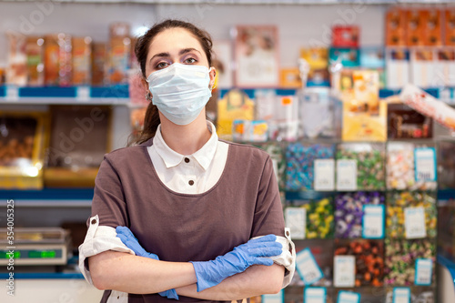 Small businesses during the pandemic.Portrait of a female worker in a medical mask and gloves posing with her arms crossed.Grocery store on the background.Concept of protection against coronavirus