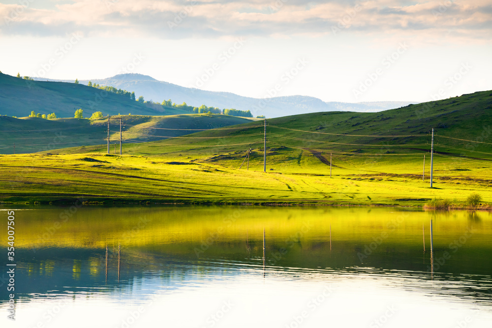 Green hills with power lines and their reflections in the lake. Beautiful summer landscape. South Ural, Russia