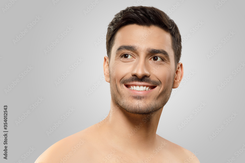 Obraz premium Young handsome man with naked shoulders and bristle, looking aside with dreamful smile, thinking of something pleasant, isolated on gray background