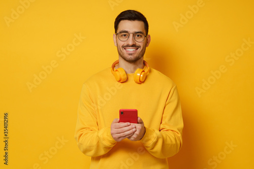 Young man in glasses, looking at camera with happy smile, holding phone in hands, wearing headphones round neck, feeling relaxed, isolated on yellow background © Damir Khabirov