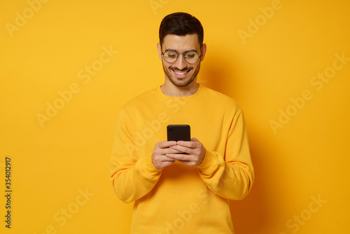 Young trendy man in glasses and sweater, feeling happy about conversation in text messages with friends via phone, smiling while looking at screen, isolated on yellow background