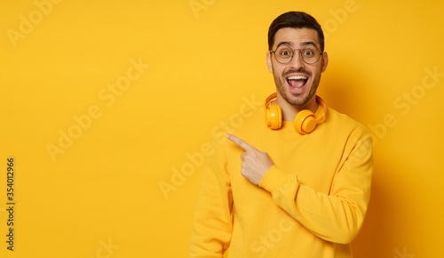 Horizontal shot of young man feeling amazed, shouting WOW, surprised by offer on copy space, pointing to it with finger, isolated on yellow background