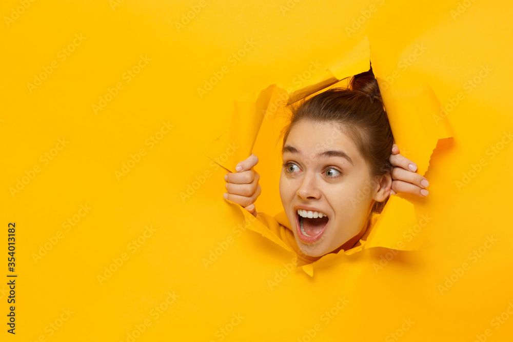 Young excited girl looking through hole in paper at beneficil commercial offer with eyes round with surprise, isolated on yellow background with copy space - obrazy, fototapety, plakaty 
