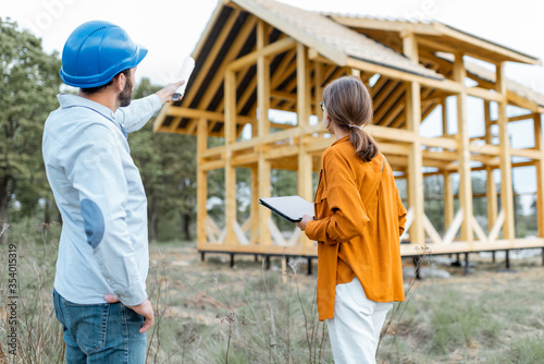 Builder with a female client near the wooden house structure on the construction site outdoors. Building and designing wooden frame house concept