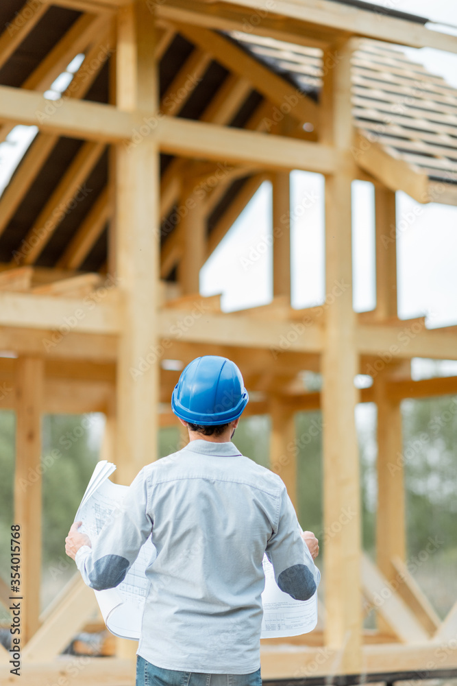 Builder or architect in hard hat supervising a project, standing with blueprints on the construction site. Building wooden frame house concept