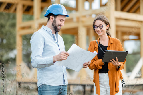 Obraz na płótnie Builder with a female client near the wooden house structure on the construction site outdoors. Building and designing wooden frame house concept w salonie