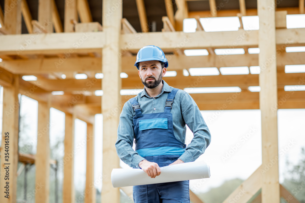 Portrait of a confident builder in blue overalls and hard hat standing with blueprints on the construction site. Building wooden frame house concept Portrait of a confident builder in blue overalls and hard hat standing with blueprints on the construction site. Building wooden frame house concept