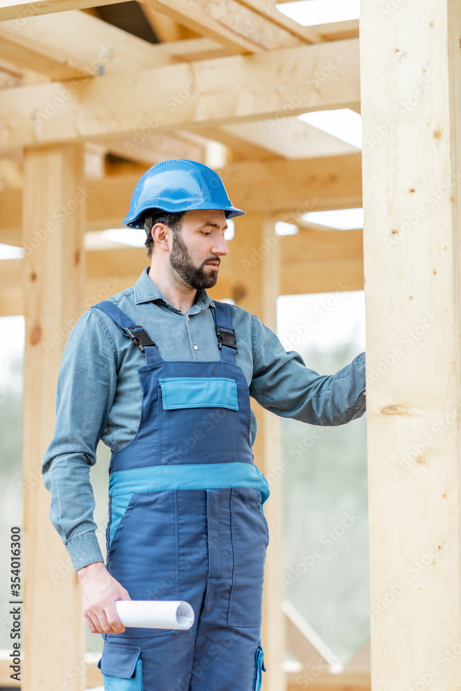 Portrait of a handsome and cofident builder in blue overalls and hard hat on the construction site. Building wooden frame house concept Portrait of a handsome and cofident builder in blue overalls and hard hat on the construction site. Building wooden frame house concept