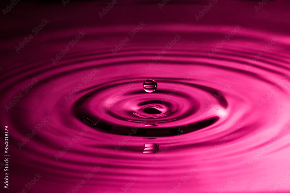 Perfect water drop splashing into smooth water causing ripples in a pink colour Perfect water drop splashing into smooth water causing ripples in a pink colour