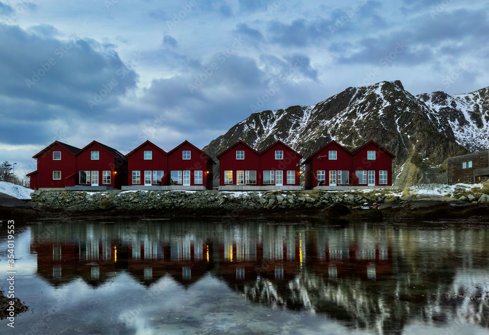 Obraz premium A horizontal shot of a symmetric row of classic red rorbu next to snowy mountain ranges reflecting water in Ballstad, Lofoten island, Norway