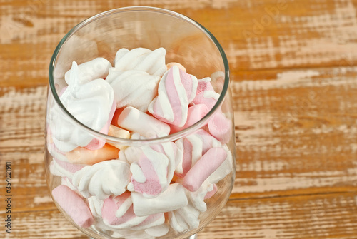 Sweets in a glass on a wooden table. Transparent utensils with marshmallows and biz on a shabby old board. Pink with white soft candies close up.