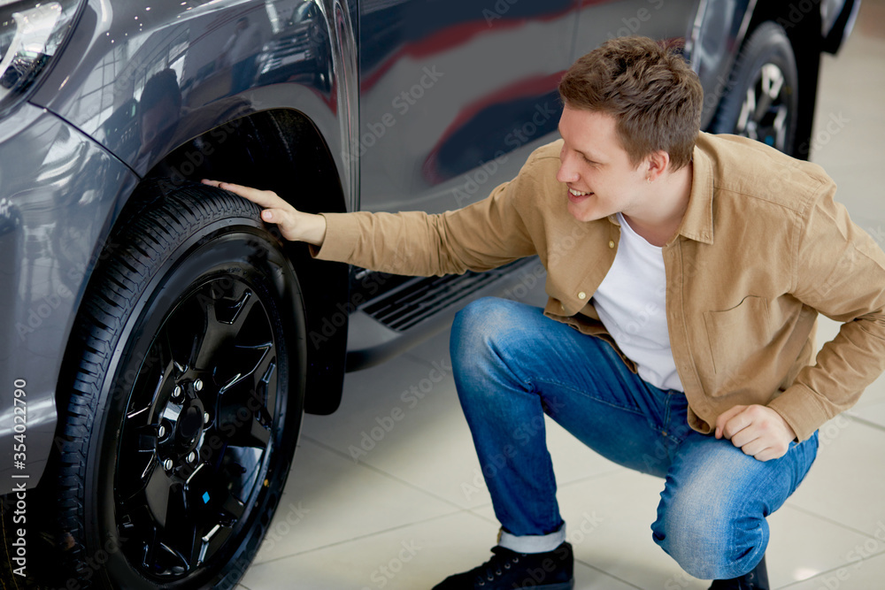 young caucasian man admires design of his new car in dealership, happy male enjoy perfectness of auto wheels