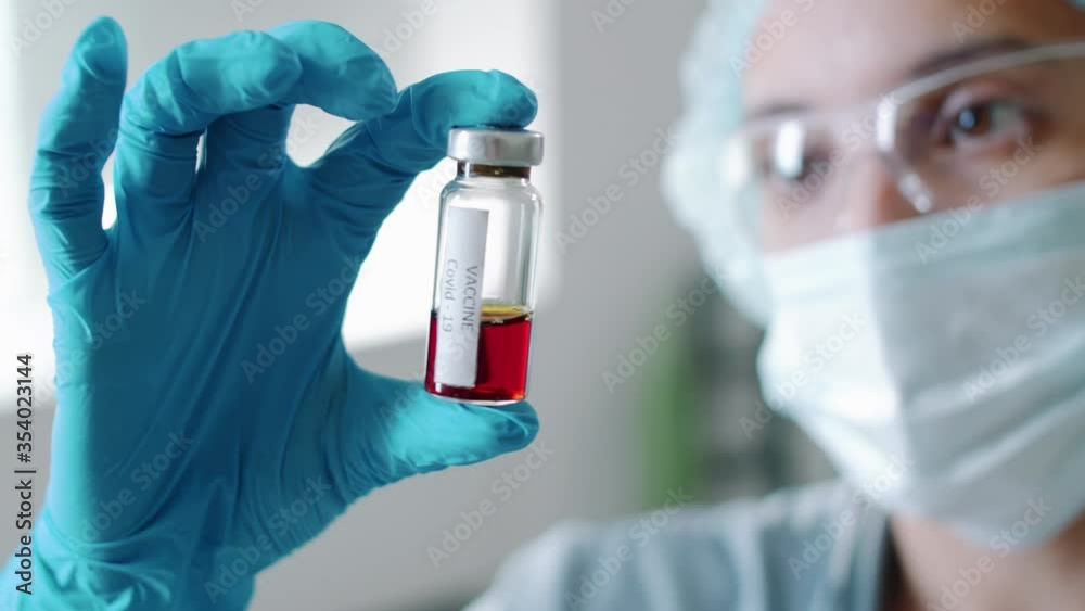 Selective focus shot of female doctor in protective gloves, mask, hat and glasses holding glass bottle with Vaccine Covid-19 inscription and examining liquid in it while working in hospital Selective focus shot of female doctor in protective gloves, mask, hat and glasses holding glass bottle with Vaccine Covid-19 inscription and examining liquid in it while working in hospital