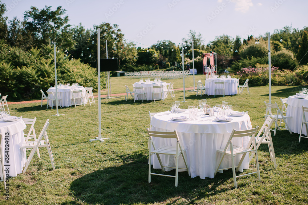 Banquet table on a green lawn. White wooden chairs. Rack and cutlery. Floral arrangement of pink flowers. On the table is a white tablecloth.