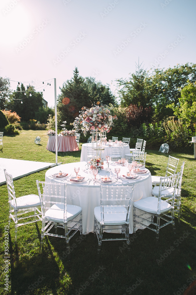 Fototapeta premium Banquet table on a green lawn. Rack and cutlery. Floral arrangement of pink flowers. Transparent banquet chairs. On the table is a white tablecloth.