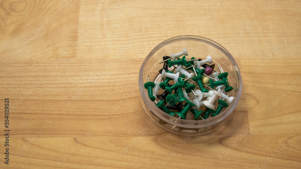 Top view of full thumb tacks in a transparent box on wooden background. It is a short nail or pin used to fasten items to a wall or board for display. Selective focus on foreground.