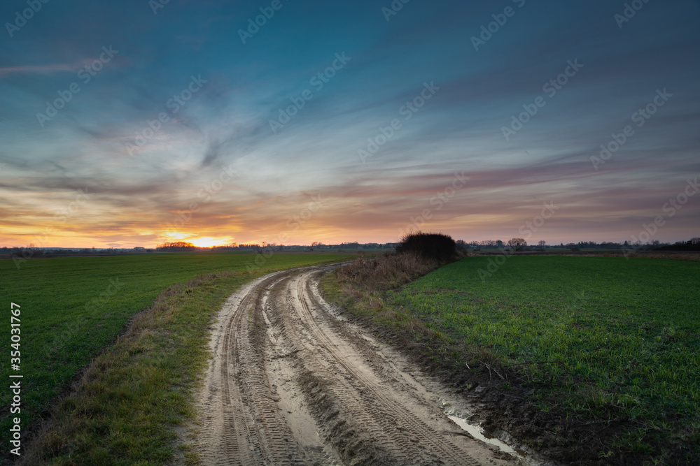 Sandy dirt road through green fields, colorful clouds on the sky after sunset Sandy dirt road through green fields, colorful clouds on the sky after sunset