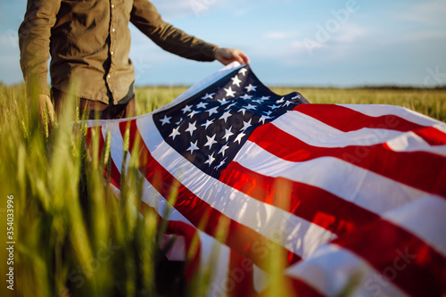 Young man wearing green shirt and cap lets the american flag fly on the wind at the green wheat field. Patriotic boy celebrates usa independence day on 4th of July with a national flag in his hands.