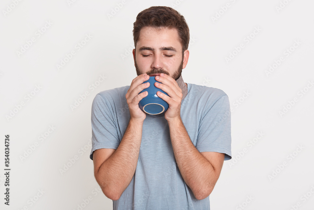 Picture of sleepy dreamy peaceful young man closing eyes, drinking coffee, posing isolated over white background in studio, wearing blue t shirt, having beard, having day off. People concept. Picture of sleepy dreamy peaceful young man closing eyes, drinking coffee, posing isolated over white background in studio, wearing blue t shirt, having beard, having day off. People concept.