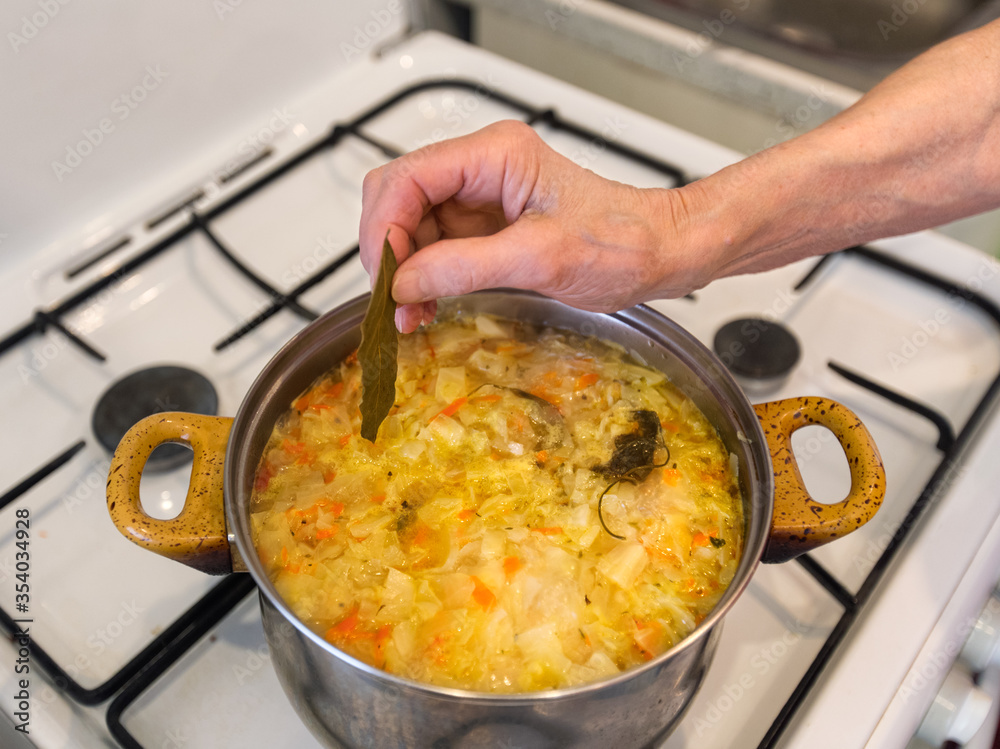 Old woman is cooking vegetable soup on gas stove. Grandmother's wrinkled hand. Pan with homemade soup on gas stove.