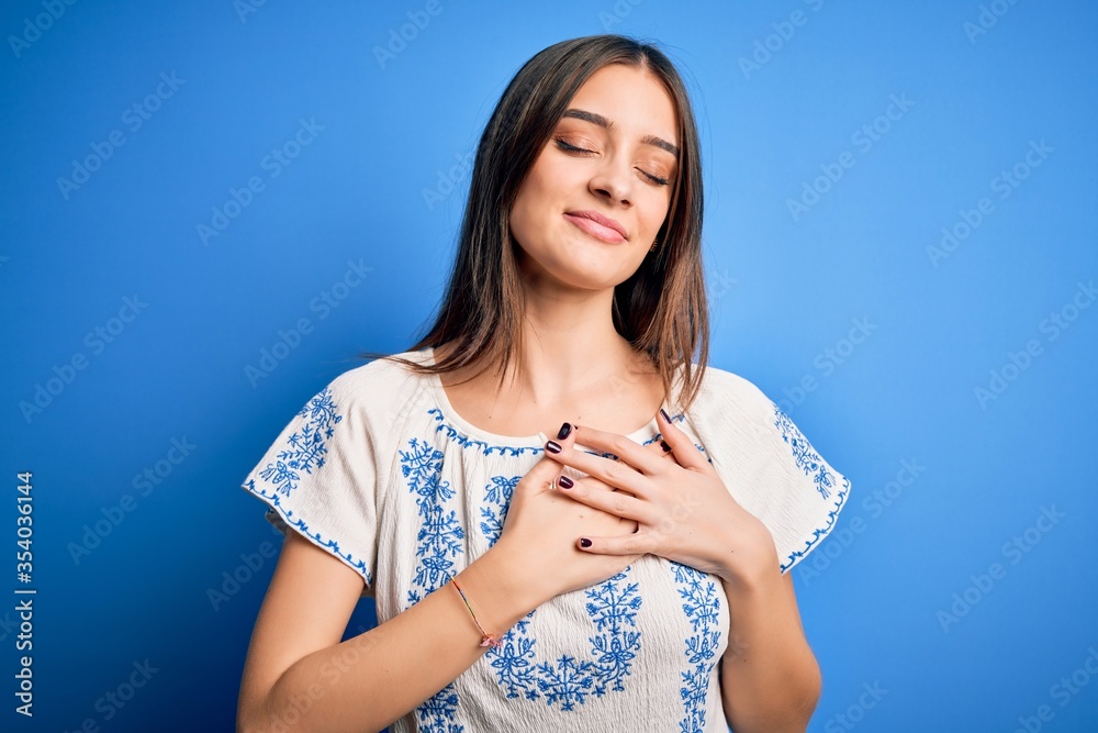 Young beautiful brunette woman wearing casual t-shirt standing over blue background smiling with hands on chest with closed eyes and grateful gesture on face. Health concept. Young beautiful brunette woman wearing casual t-shirt standing over blue background smiling with hands on chest with closed eyes and grateful gesture on face. Health concept.