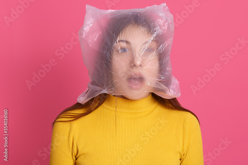 Shocked female wearing yellow shirt posing with plastic bag on her head, looks shocked and astonished, keeping mouth widely opened, stands against rose wall. photo