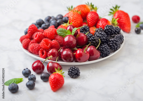 Fresh organic summer berries mix in white plate on marble background. Raspberries  strawberries  blueberries  blackberries and cherries.