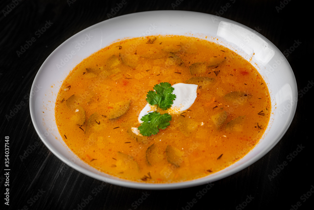 Soup with pickled cucumbers and pearl barley in white plate on black wooden table background Soup with pickled cucumbers and pearl barley in white plate on black wooden table background