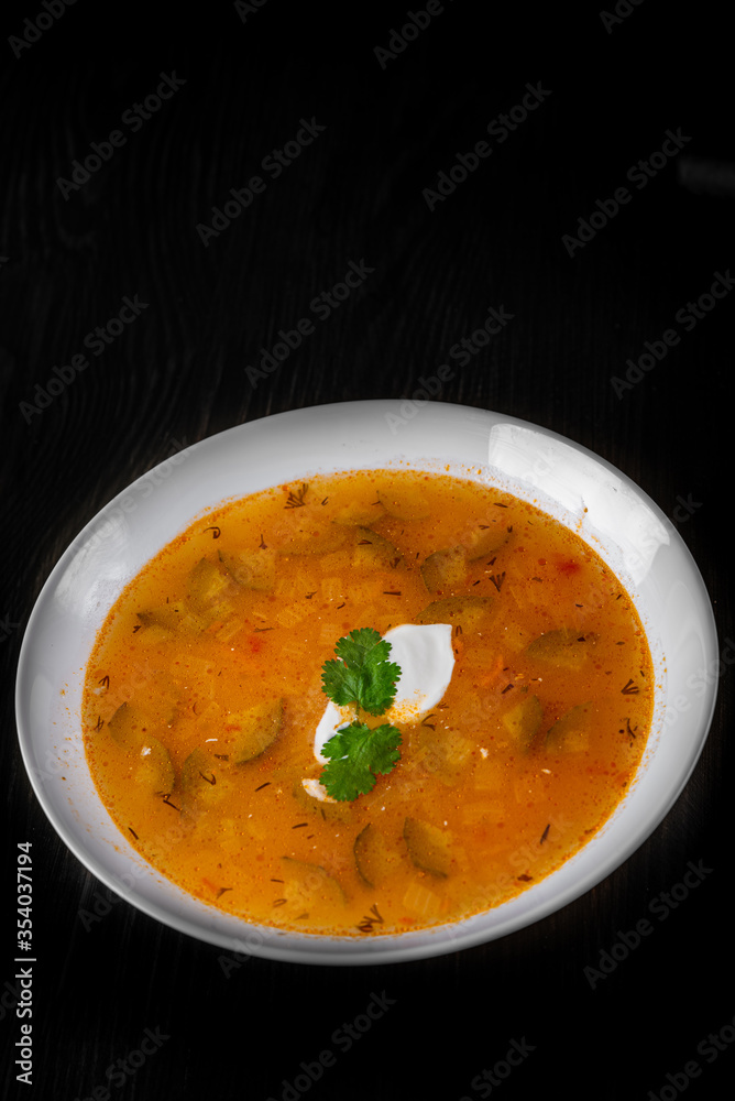 Soup with pickled cucumbers and pearl barley in white plate on black wooden table background Soup with pickled cucumbers and pearl barley in white plate on black wooden table background
