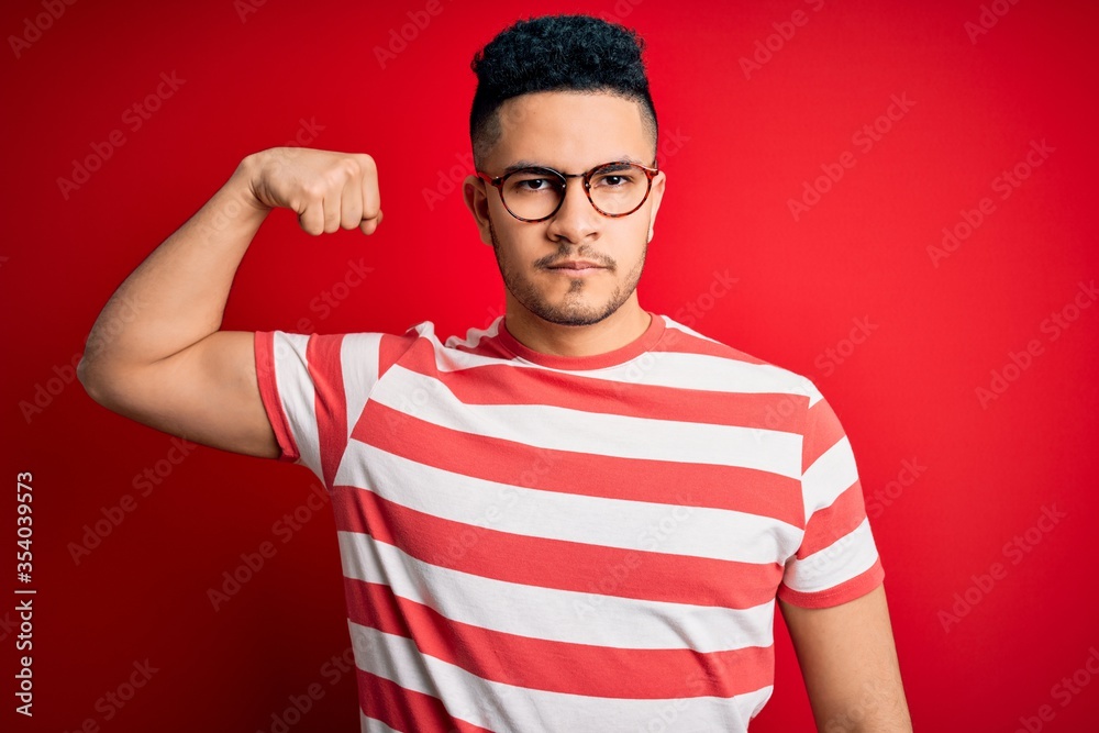 Young handsome man wearing casual striped t-shirt and glasses over isolated red background Strong person showing arm muscle, confident and proud of power