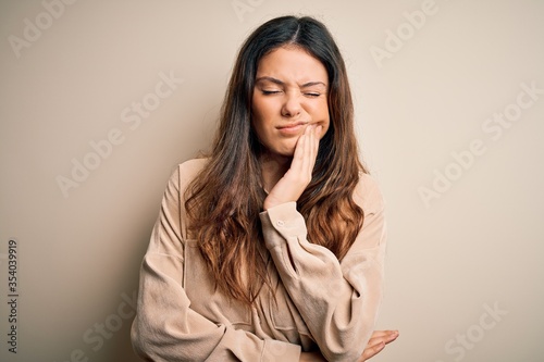 Young beautiful brunette woman wearing casual shirt standing over white background touching mouth with hand with painful expression because of toothache or dental illness on teeth. Dentist
