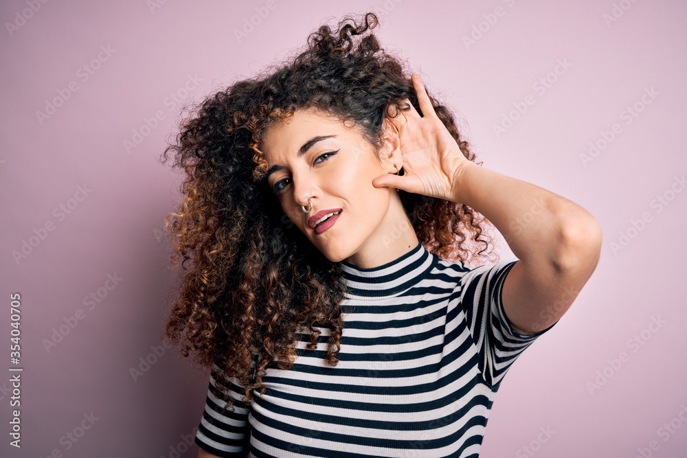 Young beautiful woman with curly hair and piercing wearing casual striped t-shirt smiling with hand over ear listening an hearing to rumor or gossip. Deafness concept. Young beautiful woman with curly hair and piercing wearing casual striped t-shirt smiling with hand over ear listening an hearing to rumor or gossip. Deafness concept.