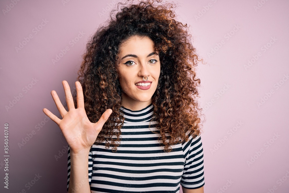Young beautiful woman with curly hair and piercing wearing casual striped t-shirt showing and pointing up with fingers number five while smiling confident and happy. Young beautiful woman with curly hair and piercing wearing casual striped t-shirt showing and pointing up with fingers number five while smiling confident and happy.